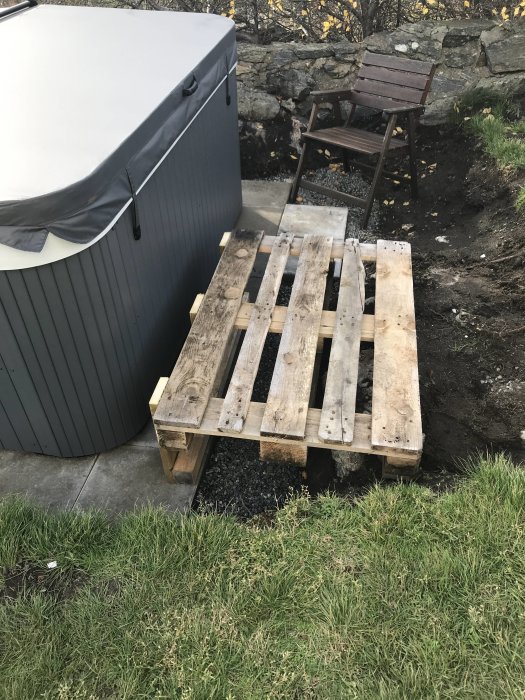 Ett spa delvis täckt av ett grått lock bredvid en oavslutad trätrallkonstruktion och en gammal stol utomhus.