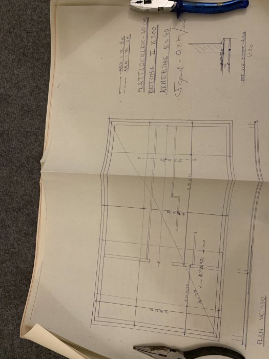 Arkitektritningar med mått och delvis skrivna anteckningar ligger på ett golv bredvid en blå häftapparat.