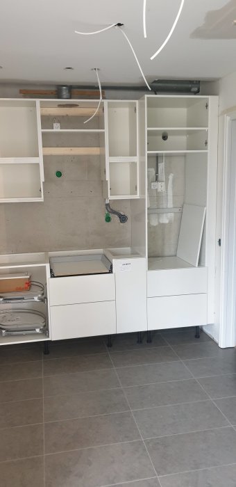 Delvis monterat vitt köksskåp utan dörrar med synliga VVS-installationer och eldragningar på betongvägg.