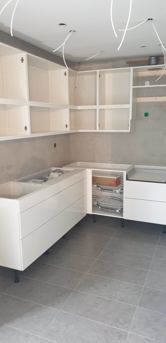 Ihopmonterade vita köksskåp och lådor i ett rum under renovering, med utsatta elledningar.