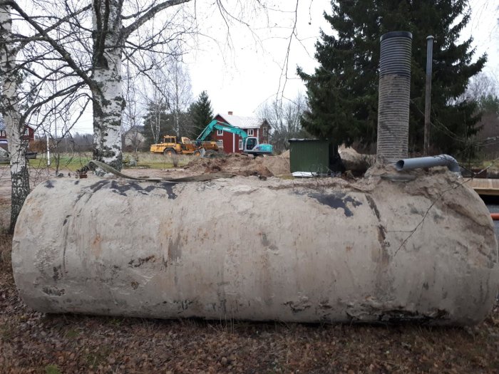 En gammal 10m3 avloppstank ligger på marken med byggmaskiner och röd stuga i bakgrunden.