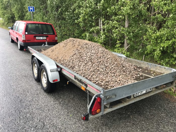 Lastbilssläp fyllt med grus parkerat bredvid en röd Volvo vid en skogskant, förberedelse för trädgårdsprojekt.