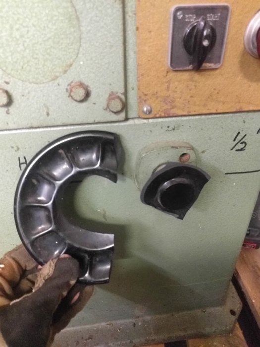 En hand i skyddshandske håller en demonterad maskindel nära en grön maskin med rattar och reglage.