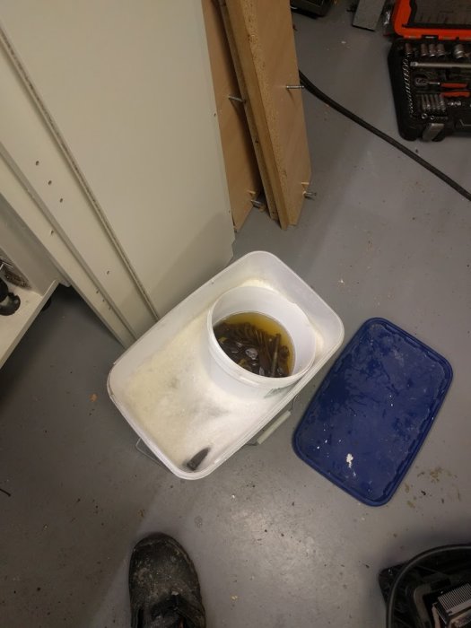 Maskindelar som ligger i blöt i en vit hink på ett verkstadsgolv, med blått lock bredvid.