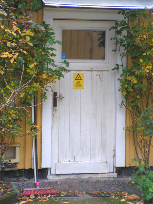 Vit panelklädd dörr på gul husfasad med varningsskylt för gasflaskor.