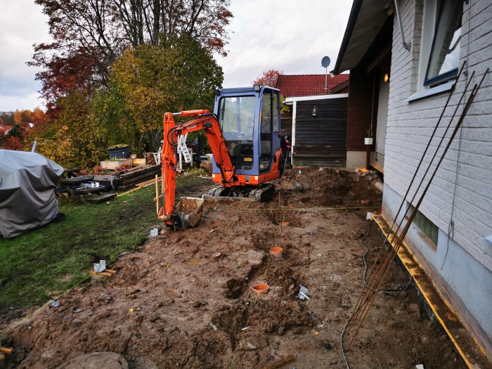 Grävmaskin vid en lerig byggarbetsplats intill ett hus, med synliga plintar och uppgrävd jord.