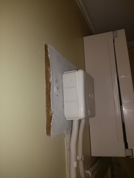 Misstänkt asbestpapp synlig bakom och under en nyinstallerad vit elcentral på väggen i ett fritidshus.