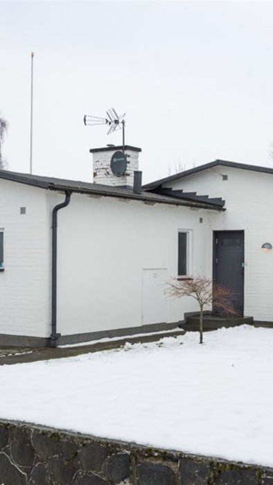 Vitputsad husfasad med skorsten där små tegelbitar trillat av, antenn nedplockad, snö i förgrunden.