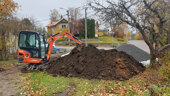 Minigrävare vid en stor jordhög vid grävning i en trädgård med höstlöv och hus i bakgrunden.
