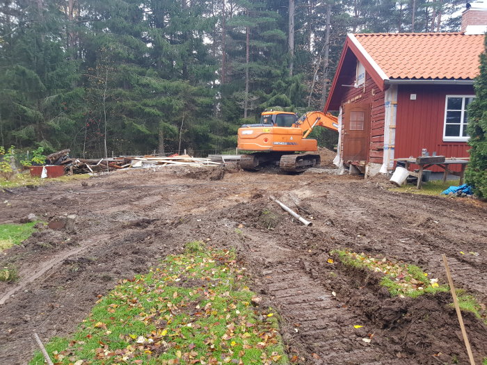 Grävmaskin vid rödfärgat hus under renovering, omgiven av uppgrovda massor och växtlighet.