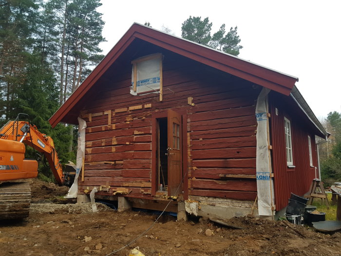 Renoveringsarbete på gammalt rött trähus med grävmaskin och uppriven grund.