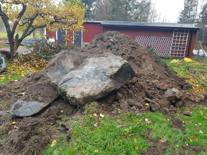 Stor sten och högar av uppgrävd jord i en trädgård, i bakgrunden syns ett rött skjul.