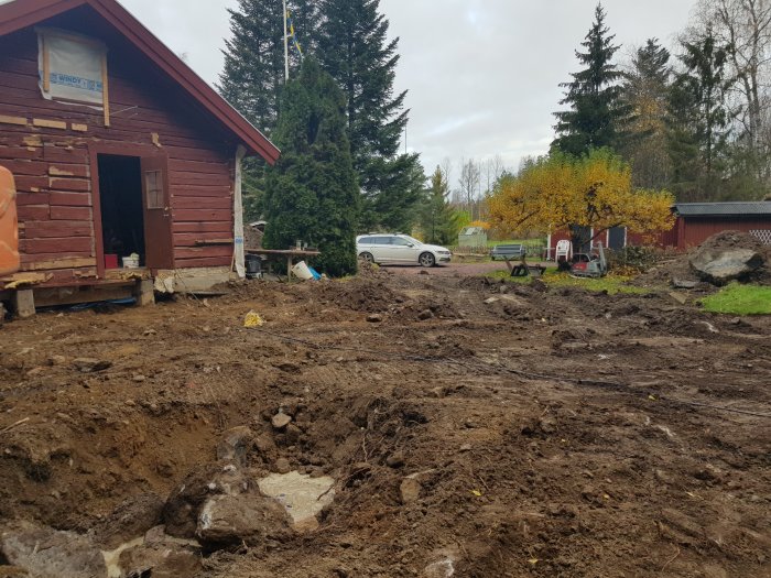 Uppgrävd mark vid rödmålat trähus under renovering med synliga stora stenar och byggavfall.