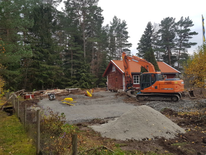 Grävmaskin vid grävarbetsplats för husgrund med röd stuga och stora stenar i bakgrunden.