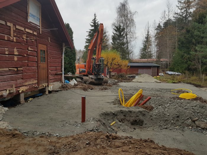 Byggarbetsplats med grävmaskin och nygjuten grund nära röd träbyggnad, gula rör synliga i marken.