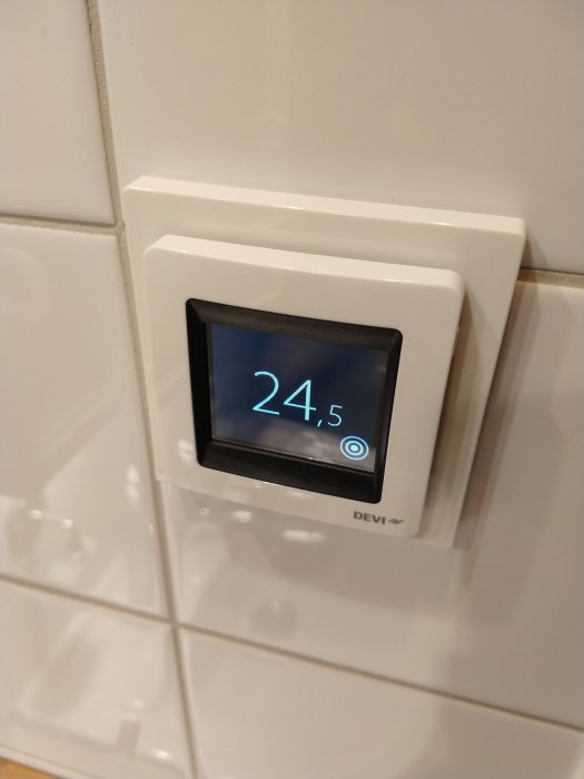 Digital termostat visande 24,5 grader installerad på vit kakelvägg.