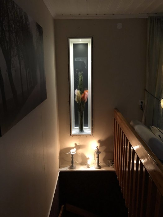 Långsmalt vertikalt fönster med smyg på en vägg bredvid en trappa, upplyst av ljus från två bordslampor.