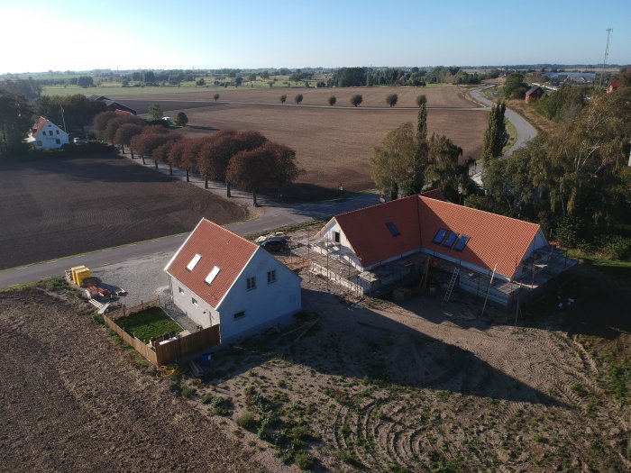 Flygbild över hus med nytt tak och pågående putsarbete, omgivet av öppna fält.