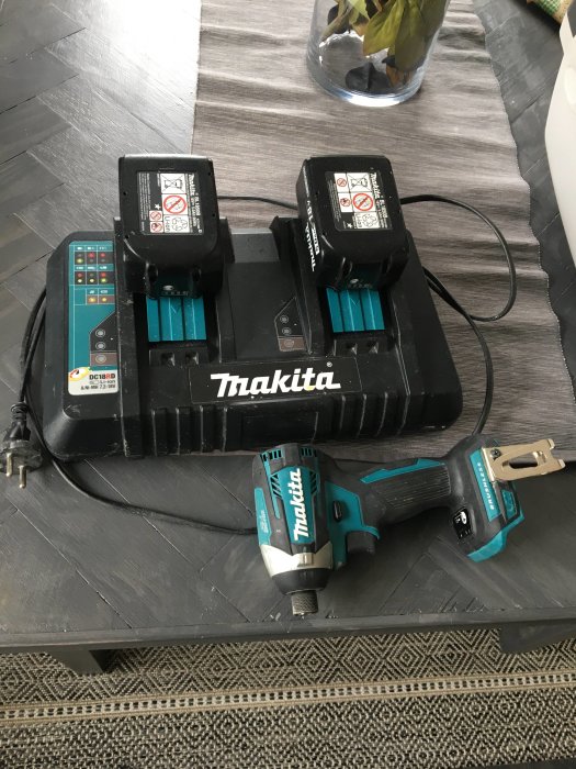 Makita dubbelladdare med två batterier i och en borr/skruvdragare på ett mörkt trägolv.