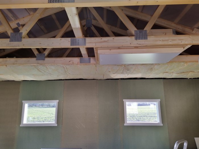 Vindslucka och isolering under takbjälkar i ett pågående byggprojekt, med synliga fönster och gråväggar.