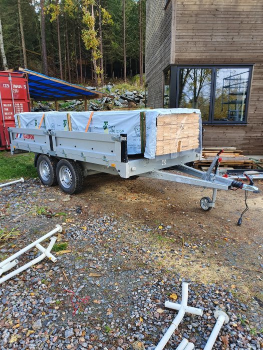 Släpvagn med paket av ytterpanel framför trähus, byggmaterial och rör på marken.