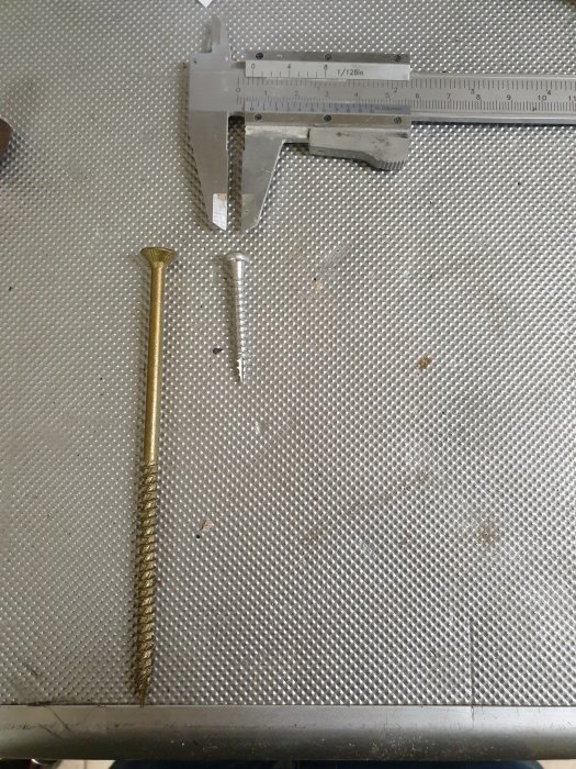 Vasemmalla messinkinen ankruuri, oikealla pienempi metalliankkuri, ylhäällä työntömitta harmaalla metallipinnalla.