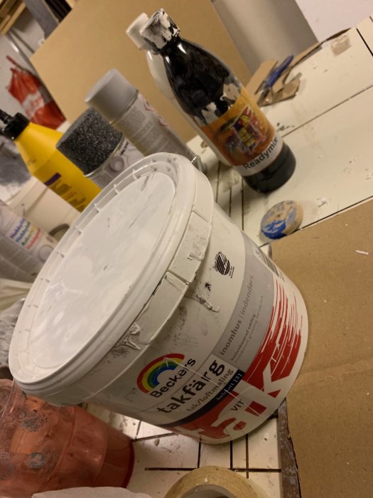 Burk med vit takfärg och svart akrylfärgflaska, använda för att måla hemmagjorda gravstenar, på en rörig arbetsyta.