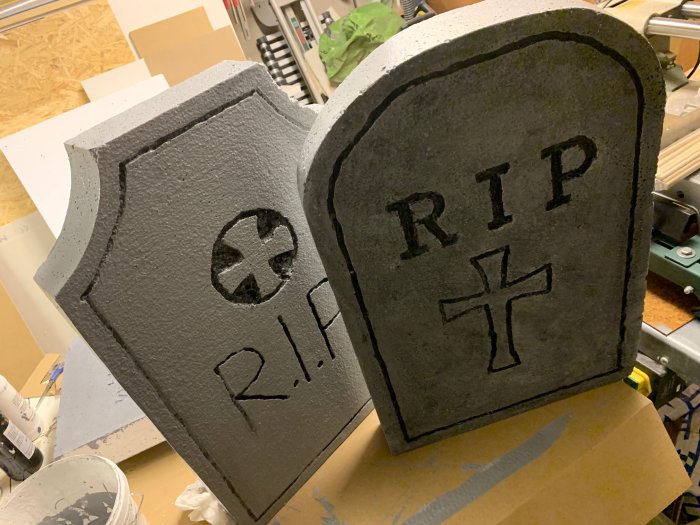 Två hemgjorda gravstenar av cellplast med inskriptionerna "R.I.P." och ett kors, står i en verkstad.