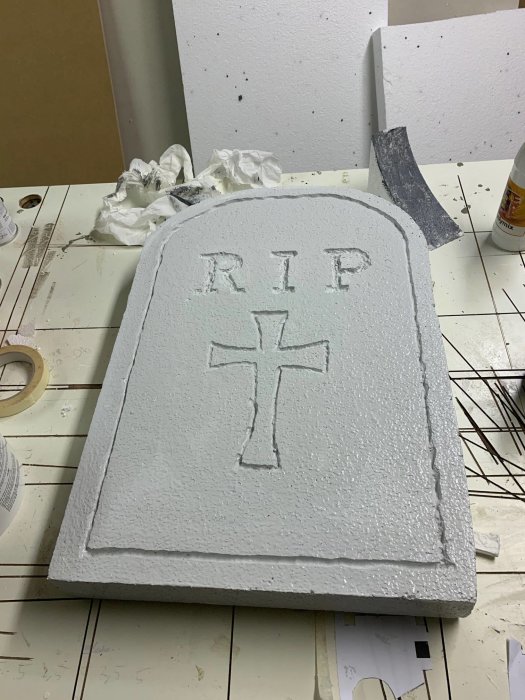 Hemgjord gravsten av cellplast med "RIP" och ett kors utskuret, i en verkstadsmiljö med målartillbehör.