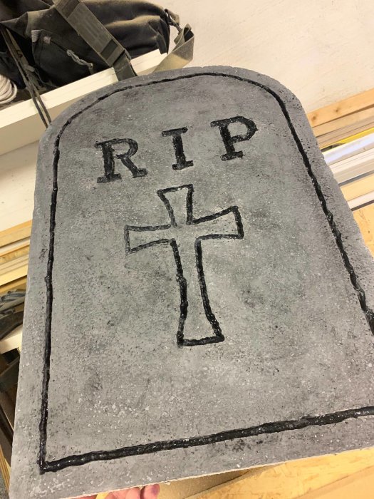 Handgjord gravsten i cellplast med inskriptionen "RIP" och ett kors, målad i grå nyanser för Halloween-decoration.