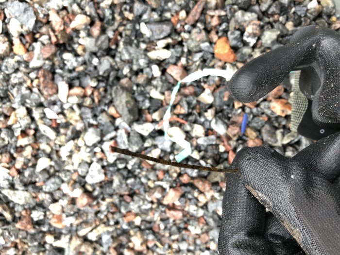 Hand i svart handske håller en tunn metallbit över grushög med orenheter som glas och plast synliga.