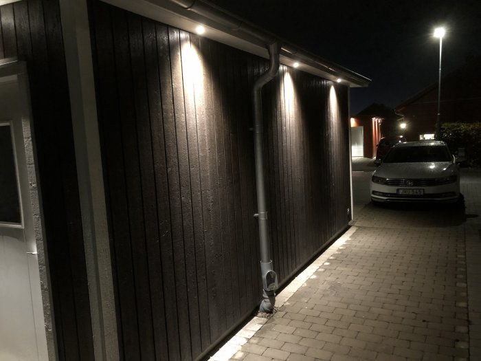 Nyinstallerad takfotsbelysning på mörkmålat hus i kvällsbelysning, med parkerad bil framför.