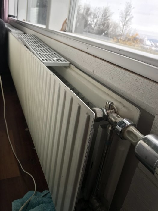 Radiator med öppen termostat vid fönster, läckage vid knopp, handduk under.