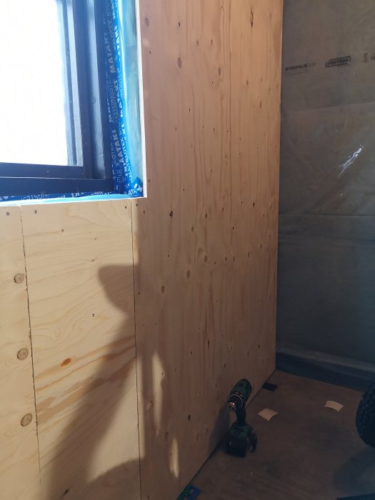 Nyinstallerad plywood på en vägg med synlig borr på golvet.