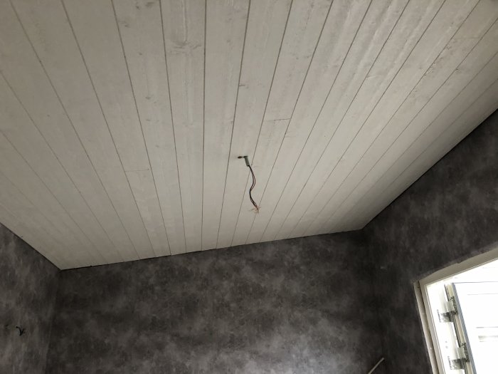 Nyinstallerat vitt träpaneltak med elektriska ledningar som sticker ut och betongmönstrad tapet på väggarna.