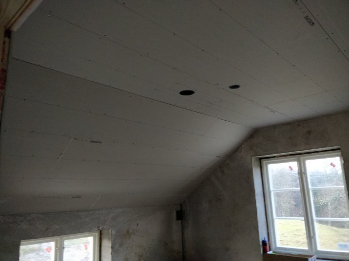 Tak i ett renoveringsprojekt gipsat med synliga skarvar och infällda spotlights, bredvid ett fönster med utsikt.
