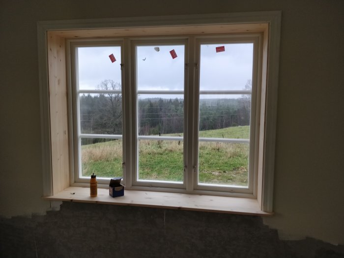 Nyinstallerat fönster med nyligen tillverkade fönstersmygar i ett rum under renovering, med utsikt över ett landskap.