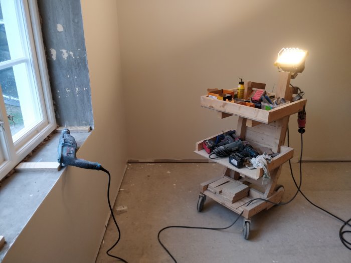 Verktygsvagn i ett renoveringsprojekt med borr, snusdosa och verktyg framför ett fönster med osparade smygar.