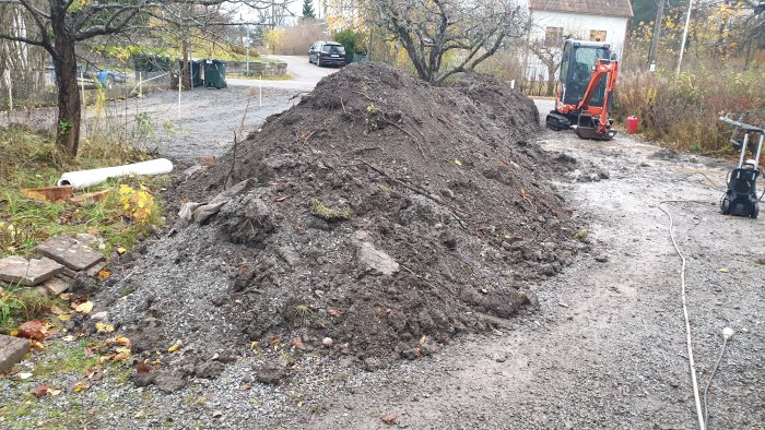 Stor hög av grävd jord och gräs som ligger på en grusdrivväg med en minigrävare i bakgrunden.