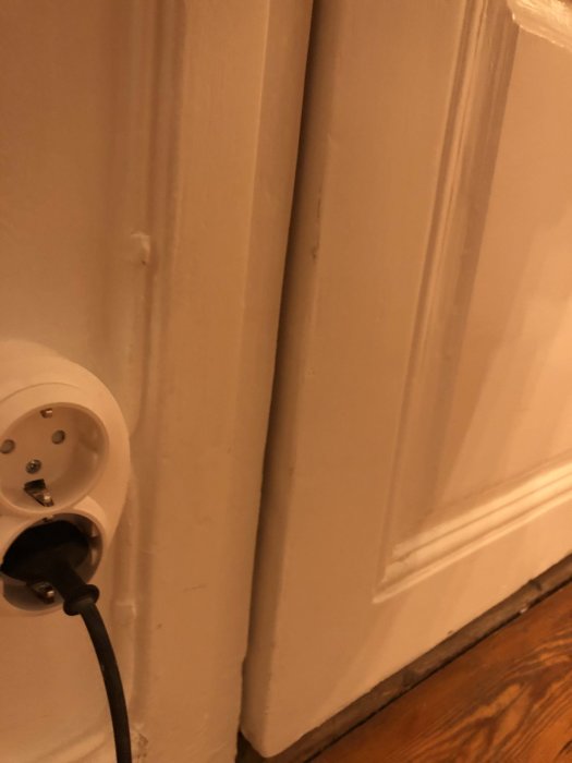 Vit dörr med skada vid nederkanten nära en eluttag och en ansluten sladd.