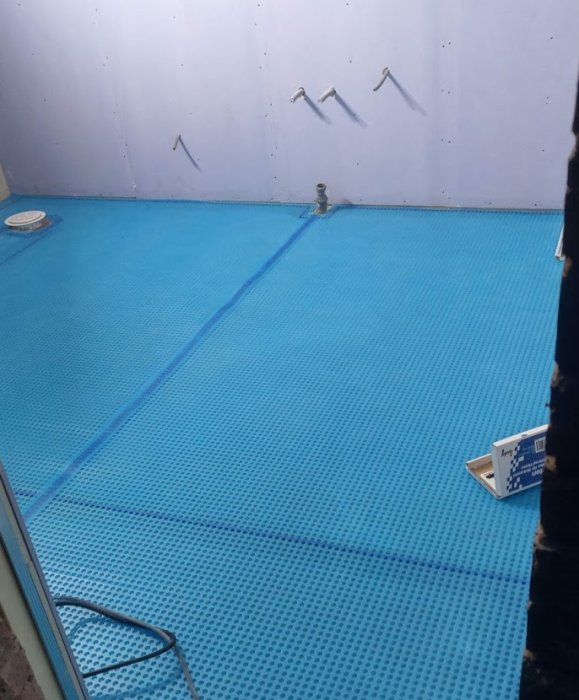 Ventilerad matta i blått lagd i ett utrymme under renovering, med synliga väggfasta rörinstallationer.
