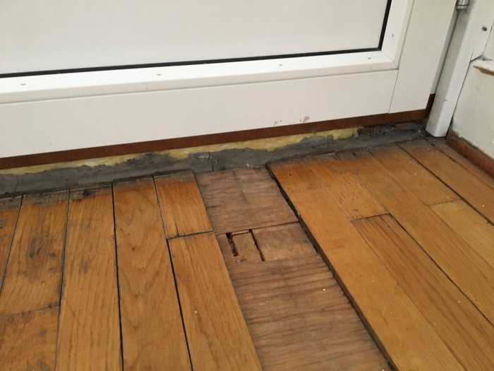 Skadad parkett där plankor släppt framför en balkongdörr, med normalt slitage på resten av golvet.