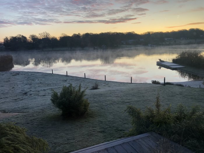 Dimmig sjö vid gryning med frosttäckt gräsmatta och brygga.