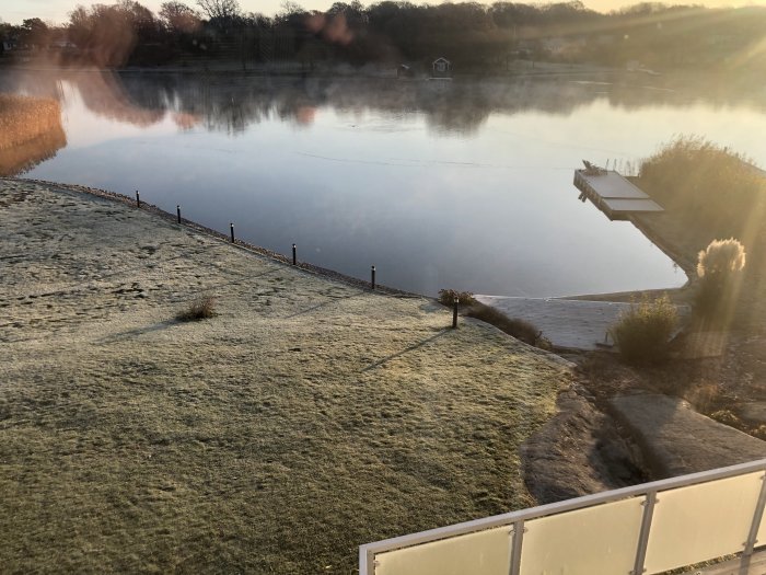 Morgonvy över en frosttäckt gräsmatta som sluttar ner mot en lugn sjö med en brygga, med solljus som speglas på vattnet.