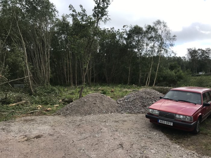 Röd bil parkerad nära två högar med grus på en plats med nyligen fällda träd och grönska i bakgrunden.