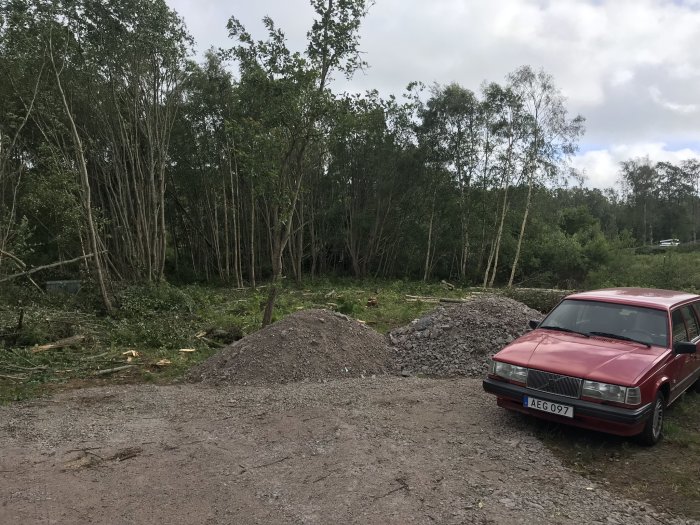 Nyfällda träd och grenar vid grushögar med en röd bil parkerad framför på en byggarbetsplats.