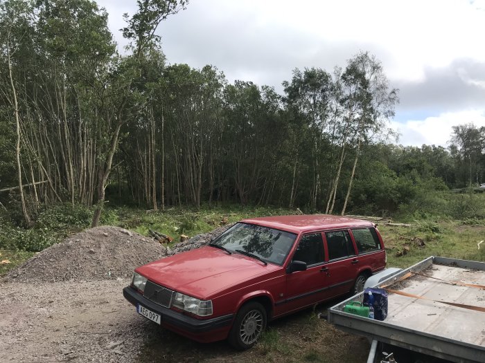 Röd kombibil parkerad vid skogskant med fällda träd och högar av grus och kvistar i bakgrunden.