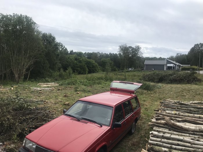 En röd bil framför fällda trädstammar och grenhögar på en avskogad yta med hus i bakgrunden