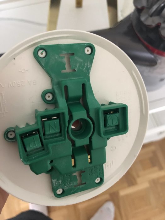 Ett öppet elektriskt takuttag med grön plastplatta och anslutningspunkt för jord, noll och fas, hållet i hand framför golv.