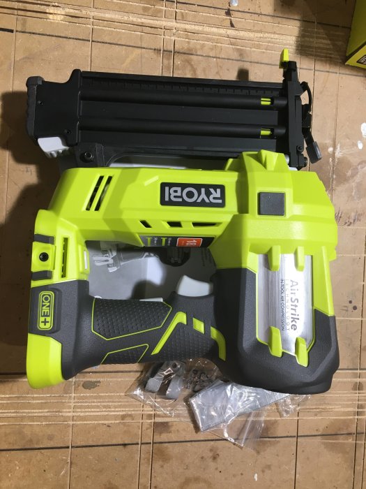 Batteridriven Ryobi AirStrike dyckertpistol på ett golv med tillbehör i en plastpåse bredvid.